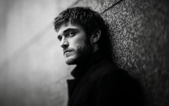 A man with short hair and a beard leans against a textured wall, gazing intensely at the camera. The black and white image creates a dramatic and moody atmosphere.