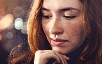 A young woman with long red hair and freckles looks down thoughtfully, resting her chin lightly on her hand. The background is softly blurred with circular light bokeh.