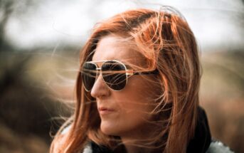 A woman with long, windblown red hair wearing aviator sunglasses looks off to the side outdoors. The background is blurred, highlighting her face and sunglasses.