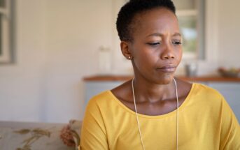 A woman wearing a yellow top sits indoors, looking down with a thoughtful or concerned expression. The background is softly lit and out of focus.