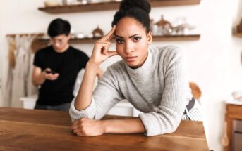 A woman in a gray sweater leans on a wooden table, looking serious and thoughtful. In the background, a man stands using his phone, slightly out of focus. The setting appears to be a cozy kitchen.