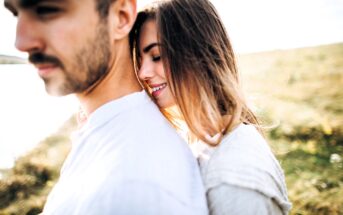 A woman with long hair leans her head on a man's shoulder, smiling with her eyes closed. The man, with short hair and a mustache, faces away from her. They stand outdoors in a sunlit, grassy field.