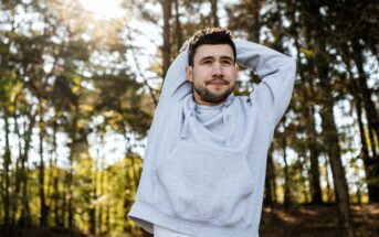 A man wearing a light gray hoodie stretches his arms over his head while standing outdoors in a sunny forest with trees in the background.