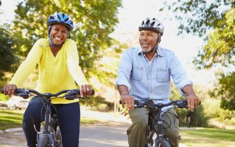 Two older adults wearing helmets, smiling and riding bicycles side by side on a sunny, tree-lined path. The woman wears a yellow sweater, and the man wears a light blue shirt.