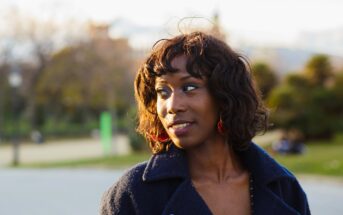 A woman with short, curly hair wearing a dark coat and red earrings stands outdoors, looking to the side and smiling, with a blurred park and trees in the background.