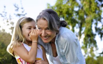 A young girl whispers into an older woman's ear, both smiling and enjoying a moment together outdoors, with green trees and sunlight in the background.