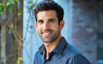 A man with short dark hair and a trimmed beard smiles at the camera. He is wearing a dark blue button-up shirt and standing outdoors next to a wall with greenery in the background.