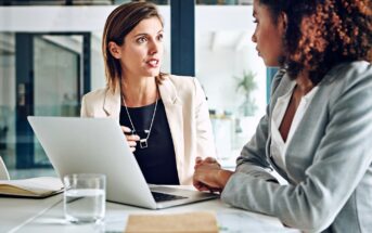 Two women in business attire sit at a desk in an office, engaged in a serious conversation. A laptop, notebook, and glass of water are on the desk in front of them.
