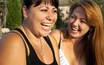 Two women are laughing together outdoors in bright sunlight. One has dark hair and wears a black tank top, while the other has long red hair and wears a striped top. Both appear happy and are enjoying each other's company.