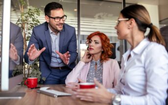 Three colleagues have a discussion at a desk in a modern office. A man in a suit gestures while talking to two women, one with red hair and the other with a ponytail, both listening attentively. Red coffee mugs are on the desk.