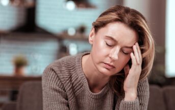 A woman with closed eyes rests her head on her hand, looking tired or stressed, wearing a knitted sweater indoors with a blurred background.