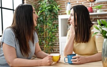 Two women sit at a table indoors, holding mugs and having an animated conversation. One woman wears a light blue top, the other a yellow one. There are plants and shelves with decor in the background.
