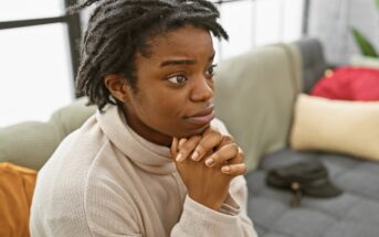 A person with short dreadlocks and a nose ring sits on a couch with their hands clasped under their chin, looking thoughtful or concerned. Pillows and a hat are visible in the background.