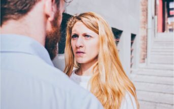 A woman with long blonde hair looks seriously at a man with a beard standing in front of her on an outdoor staircase, with sunlight illuminating her face.