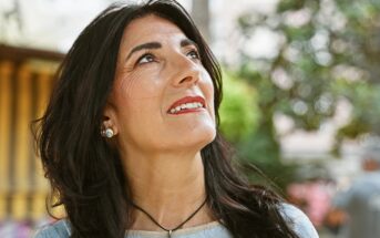 A woman with long dark hair and red lipstick looks up and smiles outdoors, with trees and blurred greenery in the background. She wears a light blue top, earrings, and a necklace.