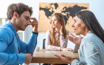 Three young professionals sit at a table in an office. One man looks stressed, touching his forehead; a woman gestures as she speaks to him; another woman in the background observes quietly. A world map is on the wall.