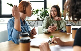 Three women sit at a table with notebooks and coffee cups, having a conversation. One woman holds a smartphone and smiles, while another gestures expressively. Sunlight streams through windows in the background.