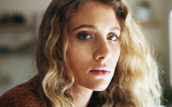 A woman with wavy blonde hair and blue eyes looks directly at the camera with a neutral expression. She is wearing a brown textured top, and the background is softly blurred.