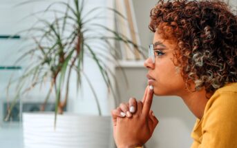 A person with curly hair and glasses sits in profile, resting their chin on their hand and appearing thoughtful. A large potted plant is in the background.