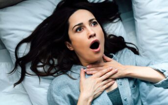 A woman lying on a bed looks alarmed, with wide eyes and mouth open, holding her hands to her throat as if struggling to breathe or feeling scared.