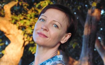 A woman with short brown hair and blue eyes smiles slightly, standing outdoors with sunlight and trees in the background. She is wearing a blue and white patterned sleeveless top.