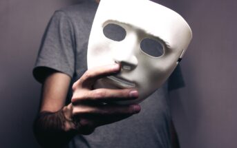 A person wearing a gray shirt holds a blank white mask in front of their chest, with only their hand and part of their upper body visible.