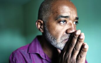 A middle-aged man with a short beard and mustache looks thoughtful and concerned, resting his hands against his face. He is wearing a purple shirt and gazing into the distance in a softly lit room.
