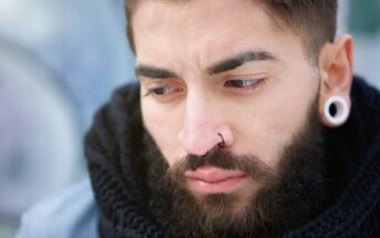 A close-up of a young man with a beard, dark hair, stretched earlobe piercings, and a nose ring, looking down with a serious or contemplative expression. He is wearing a black scarf and a blue jacket.