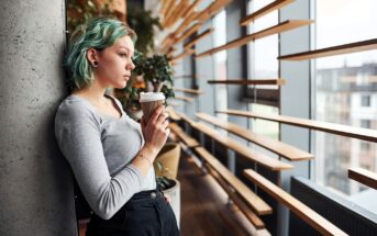 Young woman with short turquoise hair leans against a concrete wall, holding a takeaway coffee cup. She looks thoughtful, gazing out large windows in a modern, stylish space with wooden slats and indoor plants.