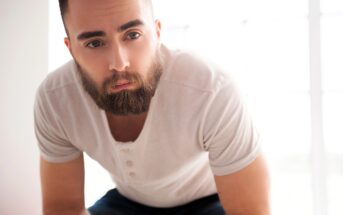 A man with a full beard and short hair wearing a light-colored henley shirt leans forward, looking directly at the camera. The background is bright and softly out of focus.