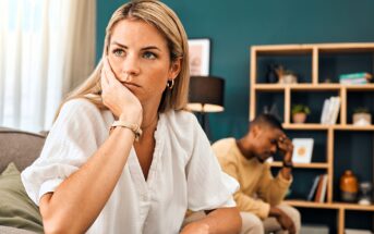 A woman sits on a couch looking away with a thoughtful, concerned expression, resting her face on her hand. In the background, a man sits with his head in his hand, appearing upset. Shelves and home decor are visible.