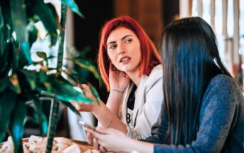 Two women are sitting indoors at a table, engaged in conversation. One has red hair and gestures while speaking; the other has long dark hair and holds a smartphone. Green plants are visible in the foreground.
