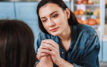 A woman in a denim shirt holds another person's hands and looks at them with a gentle, supportive expression, sitting in a kitchen with fruit visible in the background.