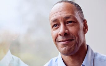 A middle-aged man with short hair and a slight smile looks thoughtfully out of a window. He is wearing a light blue shirt, and soft natural light illuminates his face.