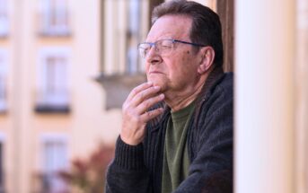 An older man with glasses and a thoughtful expression rests his hand on his chin while looking out from a balcony. He wears a dark sweater and stands against a blurred background of building windows.