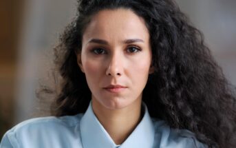 A woman with long, curly dark hair wearing a light blue collared shirt looks directly at the camera with a neutral expression. The background is softly blurred.