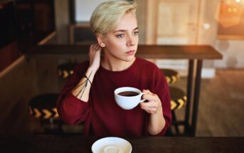 A person with short blond hair wearing a red sweater sits at a table in a café, holding a cup of coffee and looking thoughtfully into the distance. An empty saucer is on the table in front of them.