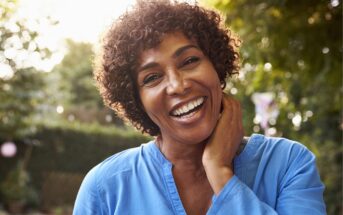 A woman with short curly hair wearing a blue top smiles warmly at the camera outdoors, with one hand gently touching her neck. The background is blurred with greenery and sunlight.