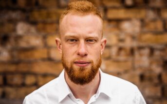 A man with short red hair and a full red beard, wearing a white collared shirt, stands in front of a rustic brick wall and looks directly at the camera with a neutral expression.