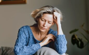 A woman with light hair wearing a blue sweater sits indoors, resting her head on one hand and looking down with a thoughtful or troubled expression.