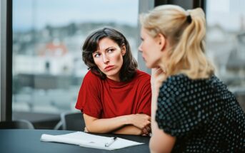 Two women sit at a table with papers and a pen. One woman, wearing a red shirt, looks skeptically at the other, who is in a black polka dot top. They appear to be having a serious conversation indoors.