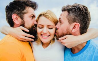 Three people cheerfully embrace. A woman in the center smiles widely, while two men on either side kiss her cheeks. They wear bright casual clothing, and the background features a clear sky.