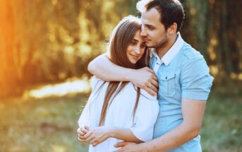 A young couple stands close together outdoors, with the man gently hugging the woman from behind. They both appear happy and content, surrounded by greenery and warm sunlight.
