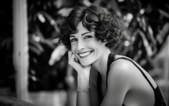 A woman with short curly hair smiles warmly at the camera, resting her chin on her hand. She is wearing a sleeveless top and a bracelet. The image is black and white with a blurred leafy background.