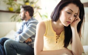 A woman sits in the foreground looking upset with her head resting on her hand, while a man sits in the background with his arms crossed, both appearing to be in the middle of an argument or disagreement.