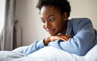 A woman in a blue hoodie rests her arms on a pillow, smiling softly and looking to the side in a bright, cozy room.