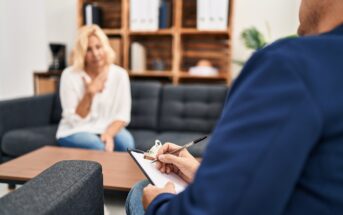 A person sits on a couch in conversation with another person holding a clipboard and pen, suggesting a therapy or counseling session in a comfortable office setting.