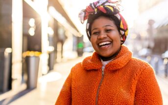 A woman wearing an orange fleece jacket and a colorful headscarf smiles brightly while standing outdoors in a sunlit urban setting.