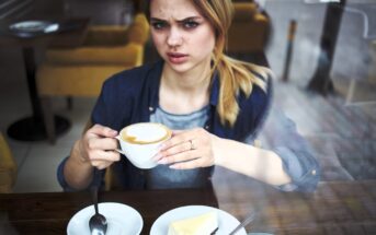 A woman sits at a café table, holding a cup of coffee and looking confused or displeased. A plate with a slice of cake, a fork, and a spoon are on the table in front of her. The photo is taken through a window.