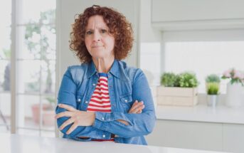 A woman with curly brown hair, wearing a denim jacket over a red and white striped shirt, stands in a bright kitchen with her arms crossed, looking serious. Green plants are visible on the counter in the background.